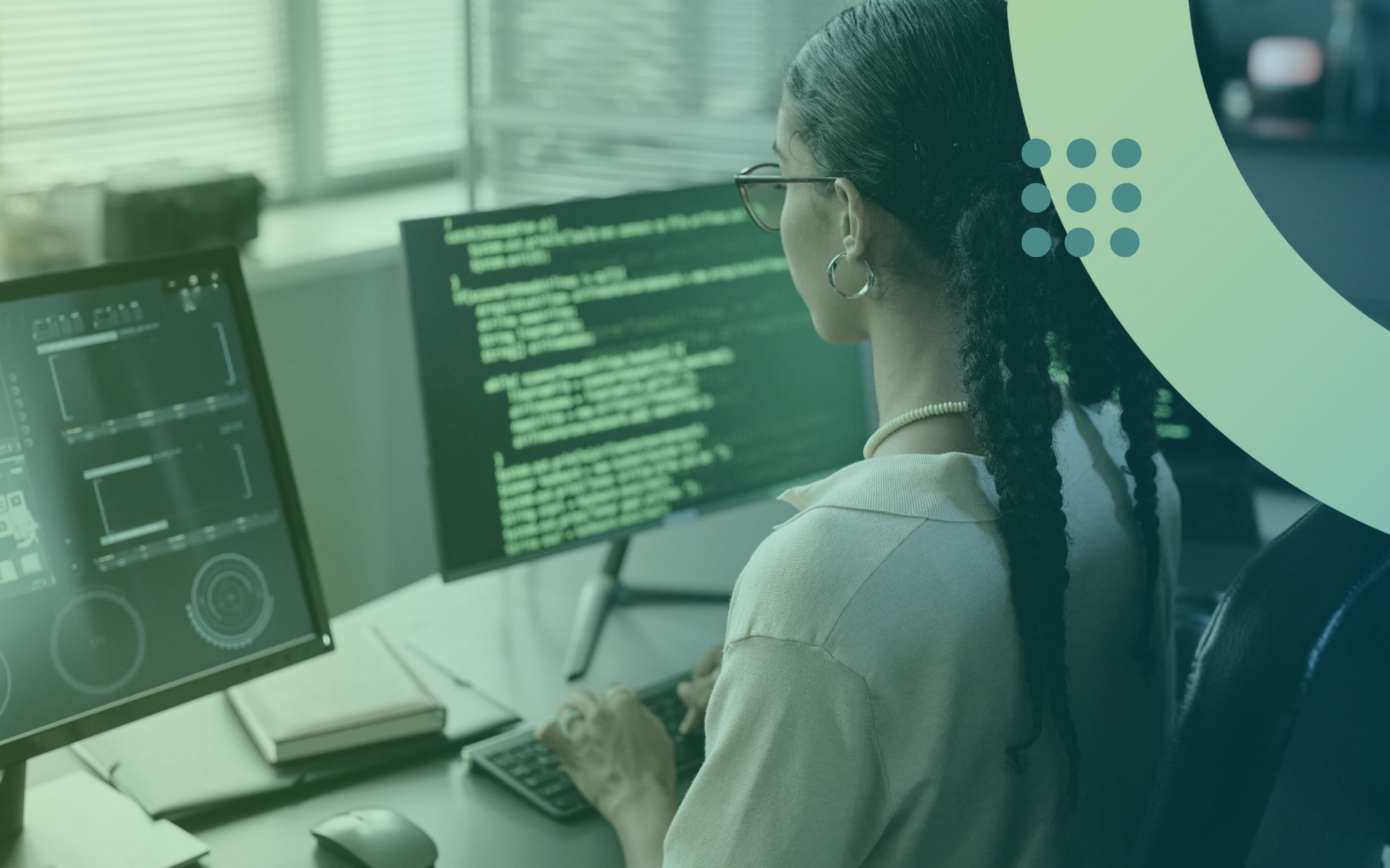 A woman with braided hair and glasses working at a desk with two computer monitors. Lines of code are visible on the screens, and her hands are positioned on the keyboard.